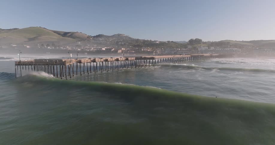 An aerial view shows a long pier extending into the ocean with waves crashing against its sides and a town visible in the background