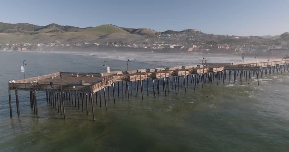 A wooden pier extending into the ocean with people walking along it under a clear sky near a coastal town and rolling hills