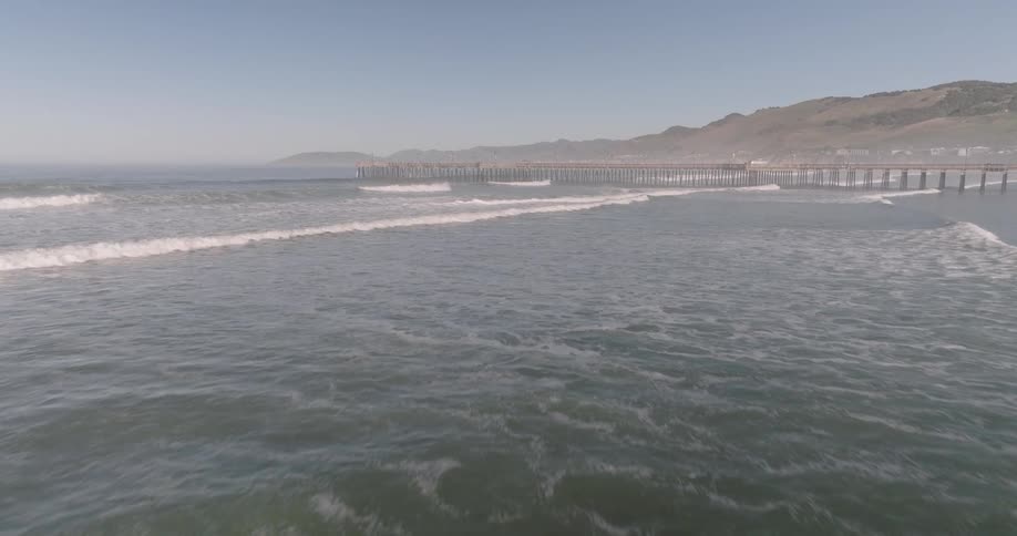 An aerial view shows waves rolling onto a sandy beach with a pier extending into the ocean and hills in the background under a hazy sky