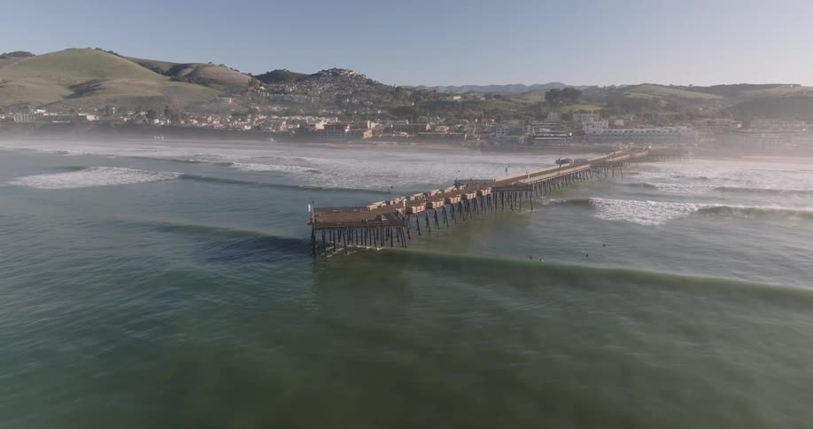 An aerial view flies away and high up from ventura pier showing the wooden structure extending into the ocean with waves and coastline