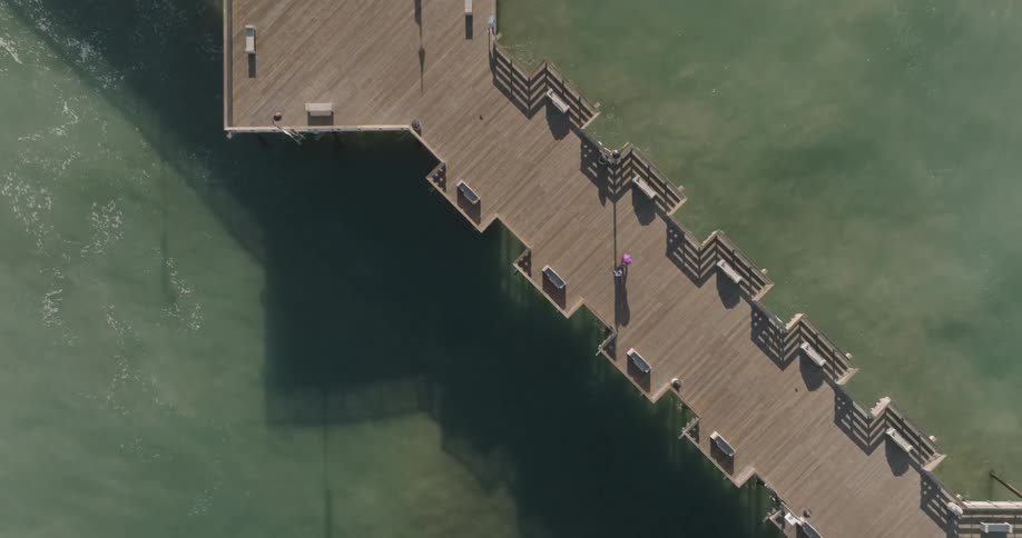 An aerial view of a wooden pier extending into the ocean with waves crashing against its sides under a bright sky and a few people wandering along its length