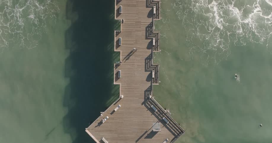 Aerial view of a wooden pier extending into the ocean with waves crashing against its sides under a bright sky