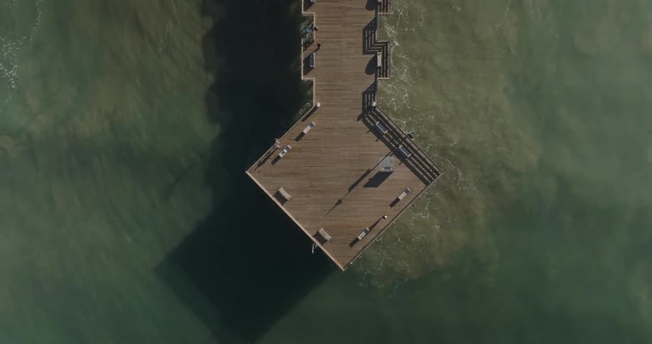 An aerial view shows a wooden pier extending into the ocean with waves crashing against its sides under a bright sky