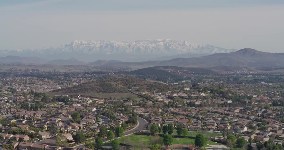 An aerial view of a suburban neighborhood with closely packed houses and trees nestled against a backdrop of hazy mountains with snow capped peaks