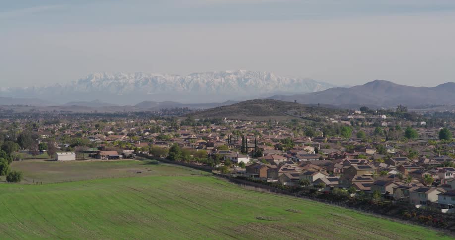 An aerial view of a suburban neighborhood with closely packed houses and trees nestled against a backdrop of hazy mountains with snow capped peaks