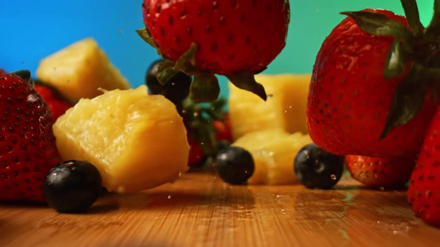 A closeup shows strawberries blueberries and chunks of pineapple being dropped onto a wooden surface the view pulls back