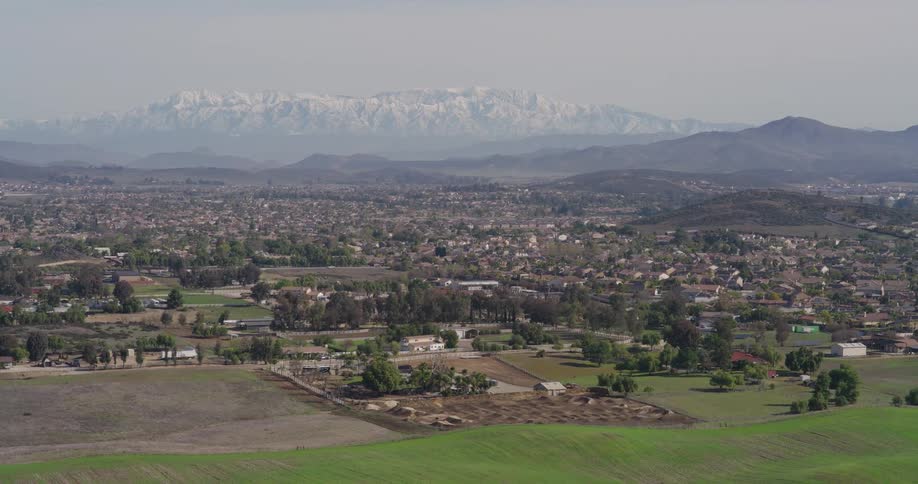 An aerial view shows a sprawling suburban landscape with houses and trees leading to a mountainous backdrop with snow capped peaks under a hazy sky