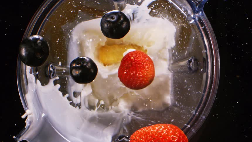 Strawberries a plum and pineapple pieces fall into a blender splashing milk as they blend together