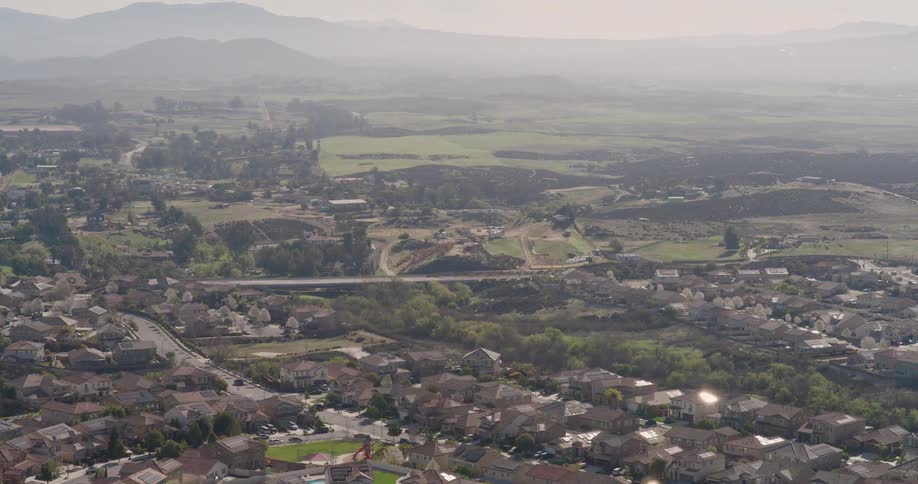 An aerial view shows a suburban neighborhood nestled amongst rolling hills and a hazy landscape