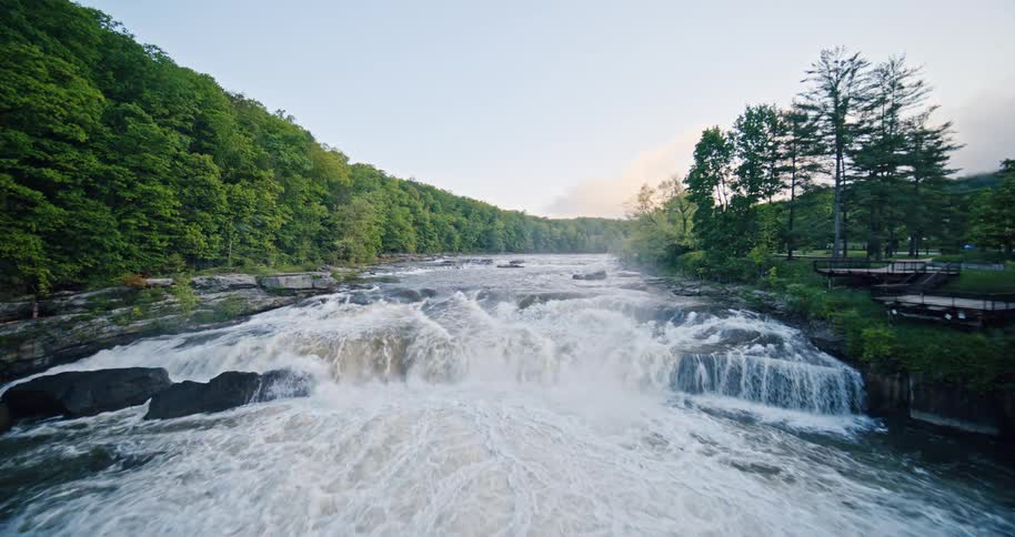 A wide rushing river flows over rocks forming a series of small waterfalls framed by lush green forested hills