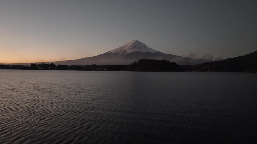An aerial perspective approaches mount fuji its iconic snowcovered peak distinctly visible against a calm twilight sky positioned rising serenely beyond a wide dark lake