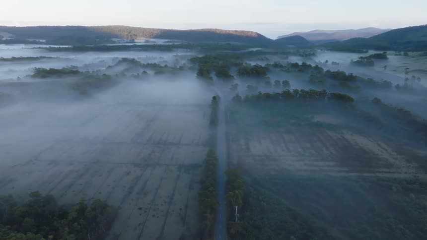 An aerial view shows a foggy valley with forested hills in the distance