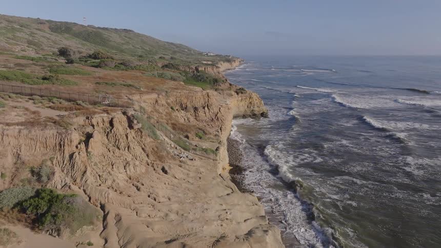 An aerial view flies forward above a dramatic rugged coastline showcasing cliffs meeting the ocean with waves crashing against the shore