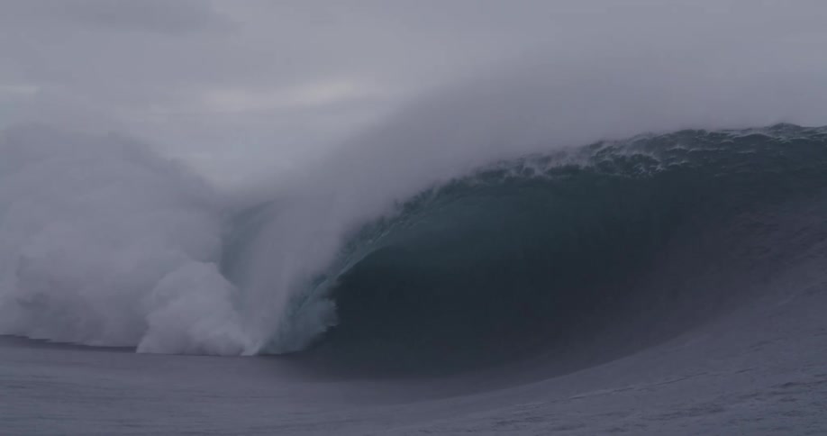 A large wave crashes in the ocean under a cloudy sky
