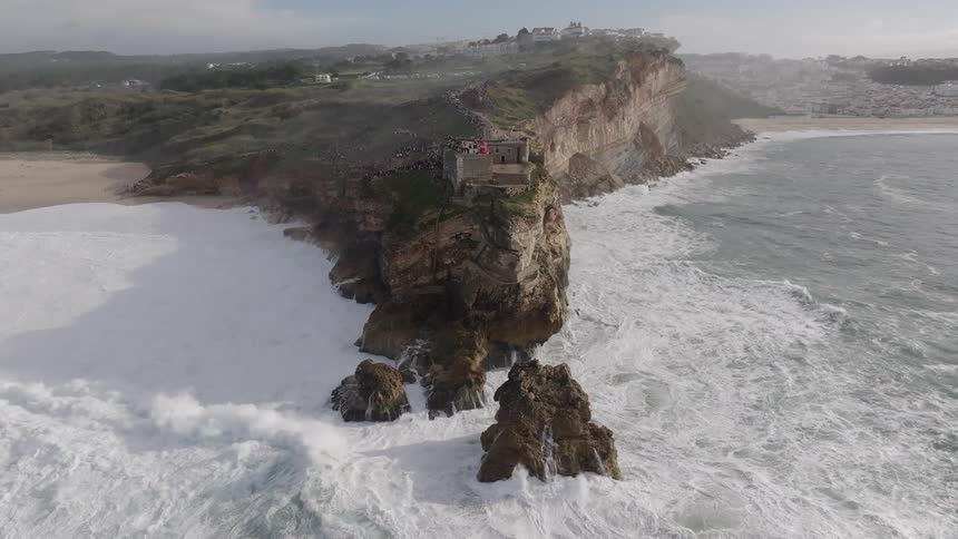 An aerial view captures the forte de so miguel arcanjo in nazar portugal on a cliff with nearby people overlooking strong waves breaking into white against rocky formations