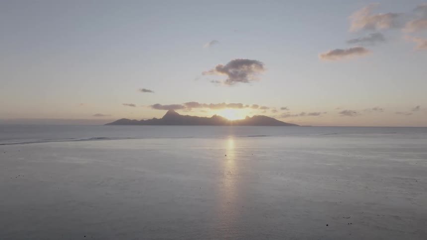 A distant island silhouette is centered in a wide shot of a calm sea at dusk with the setting sun reflecting on the waters surface