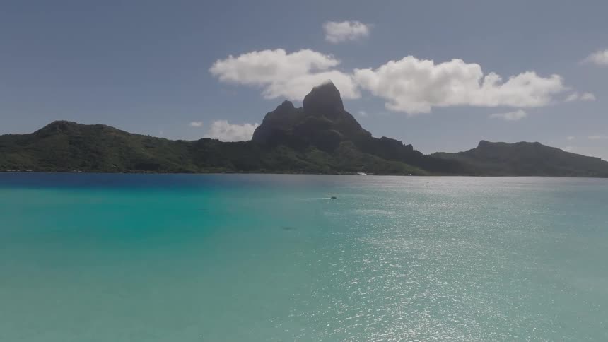 An aerial view flies over the stunning turquoise waters of bora bora with a distant sailboat towards a majestic cloudtopped mountain peak