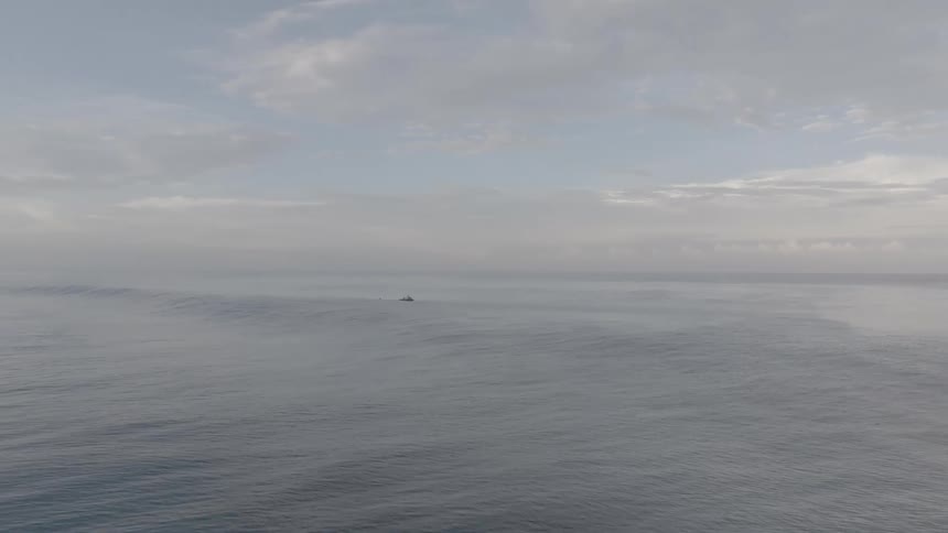 An aerial view shows calm ocean waters with surfers scattered across the surface stretching toward a muted horizon