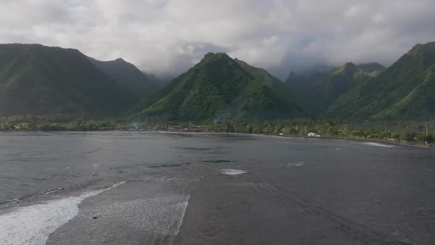 An aerial view flies toward a lush mountainous coastline with rippling waves approaching the shore under a cloudy sky