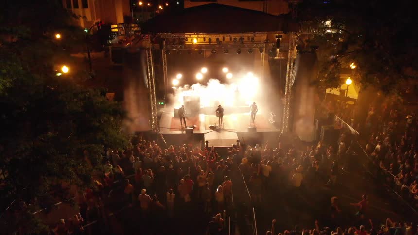 A nighttime outdoor concert features a performer on a brightly lit stage surrounded by a large crowd gathered on the streets
