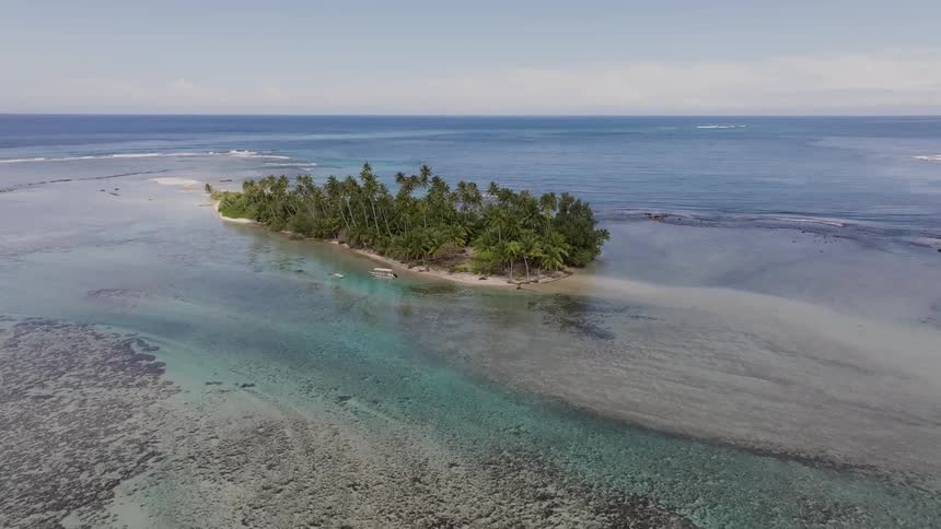 An aerial of a small tree covered island surrounded by clear shallow water with visible coral reefs and a boat is floating nearby