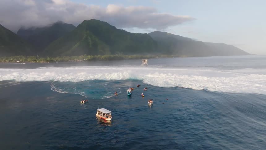An aerial view shows a large wave breaking in the ocean with several boats and people scattered nearby against a backdrop of lush green mountains