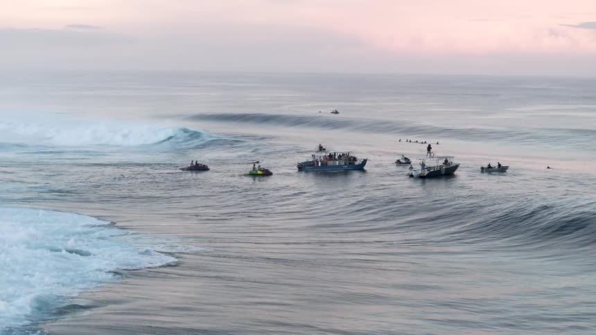 Large waves crash in the ocean with several boats and surfers visible in the distance
