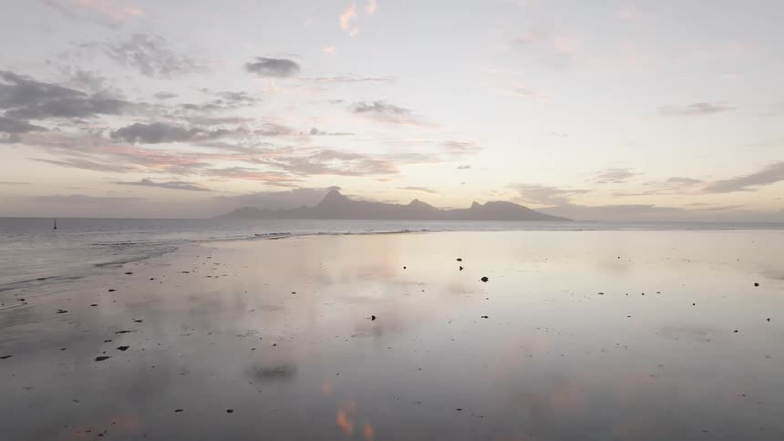 A wide reflective beach stretches to the horizon with distant mountains under a cloudy sky at dusk