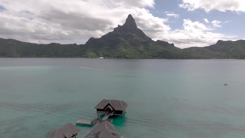 An aerial view moves over luxurious overwater bungalows in bora bora with a boat in the clear water heading towards the iconic mount otemanu