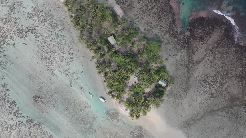 An aerial view starts low rises above a small tropical island with huts and trees surrounded by a coral reef in shallow turquoise water