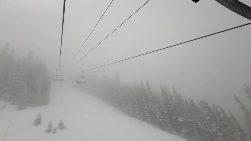 A view from a ski lift ascending a snowy mountain in foggy conditions showing empty chairs descending on the other side amongst pine trees