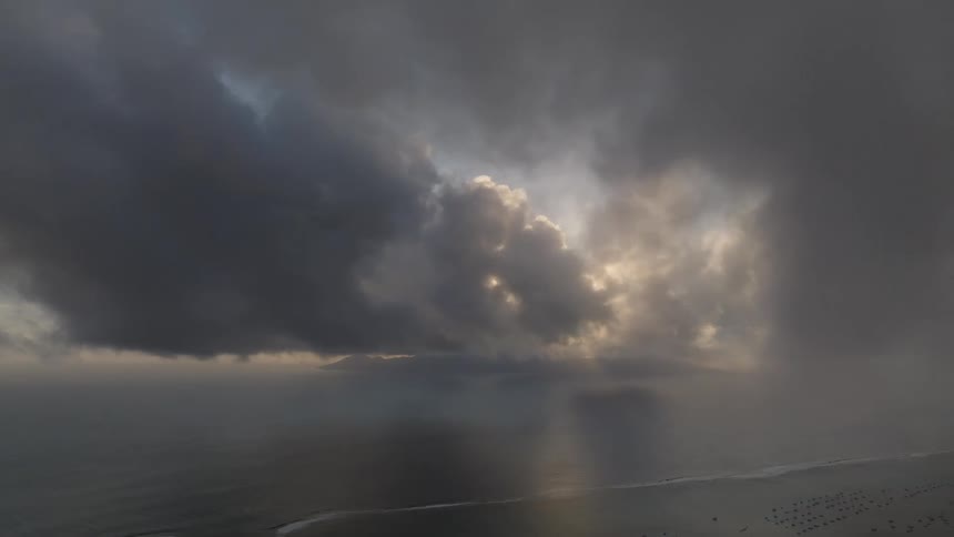 Dark dramatic clouds fill the sky above a calm sea with sunlight breaking through in patches