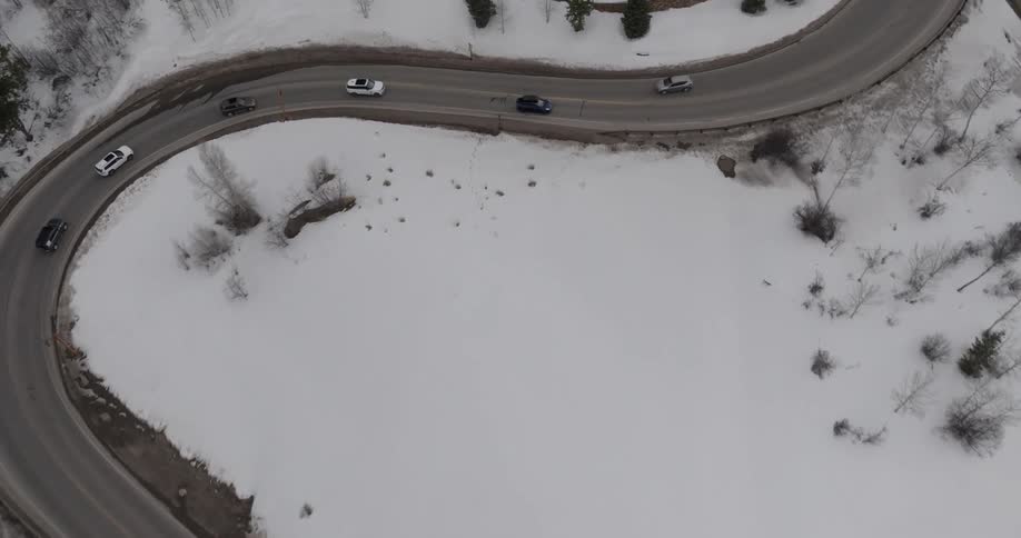 An aerial view of a winding road cutting through a snowy landscape with several cars traveling along it