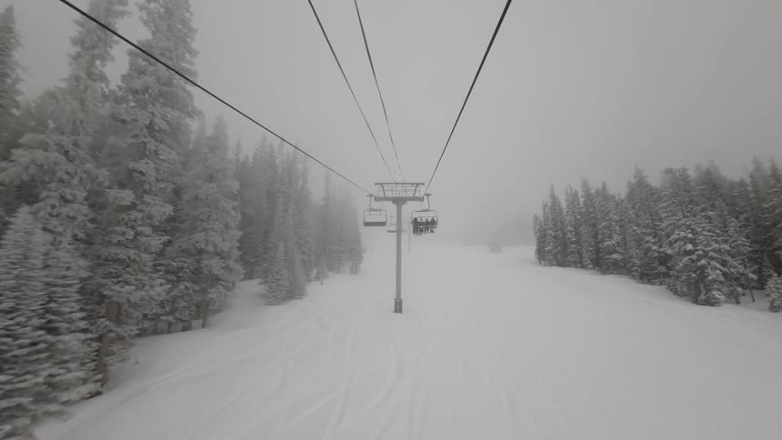 A view from a ski lift ascending a snowy mountain showing people on the chair ahead and empty chairs descending on the opposite side