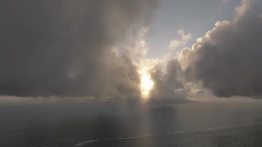 The sun shines from behind an island at sunrise while waves crash on the beach on a cloudy day