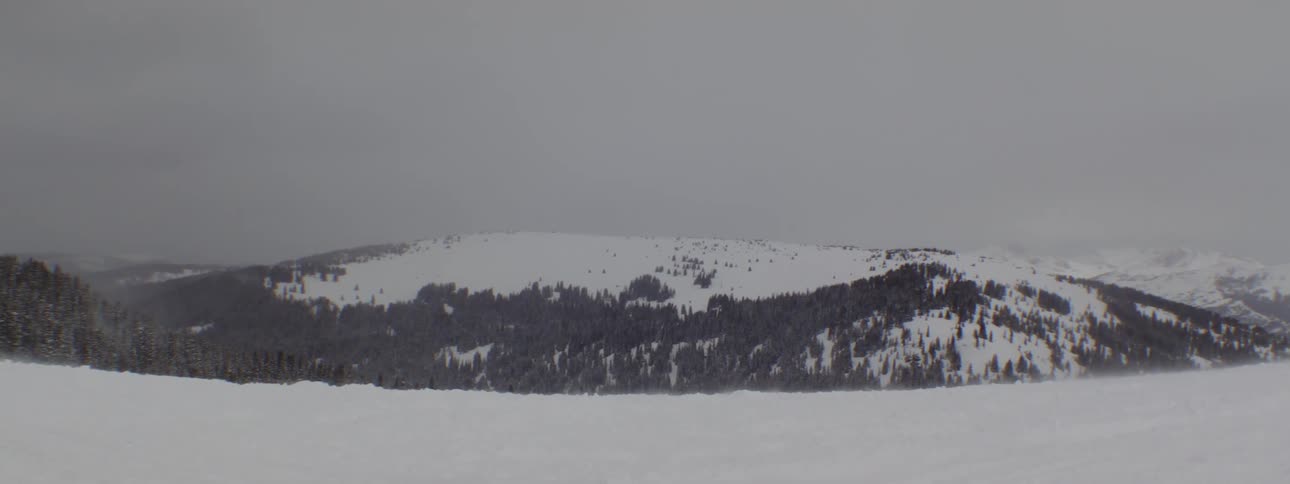 A wintry mountain shows strong winds sweeping across snowy scenery lifting snow into the air creating a white texture under a soft and cloudy grey expanse presenting a wild wintry landscape