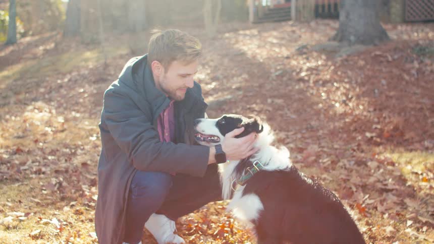 Crouching man smiles while petting a dog on an autumn day near a rural home