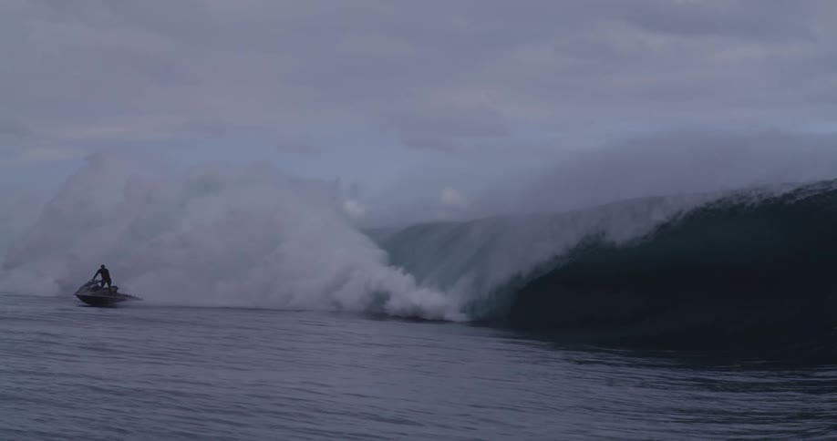 A large wave crashes in the ocean under a cloudy sky with a jet ski visible in the foreground