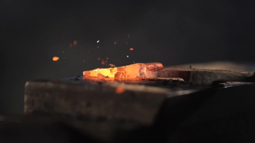 A hammer strikes a glowing hot piece of metal on an anvil sending sparks flying in a blacksmithing process