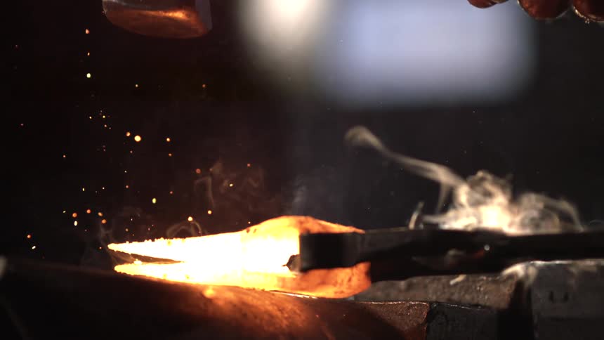 A blacksmiths hand holds a hammer striking a glowing hot piece of metal on an anvil with sparks flying