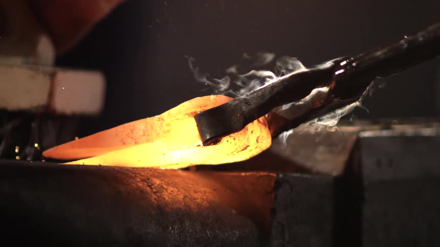 A glowing heated piece of metal is held by tongs while a metal wire brush scrapes over it against a dark background