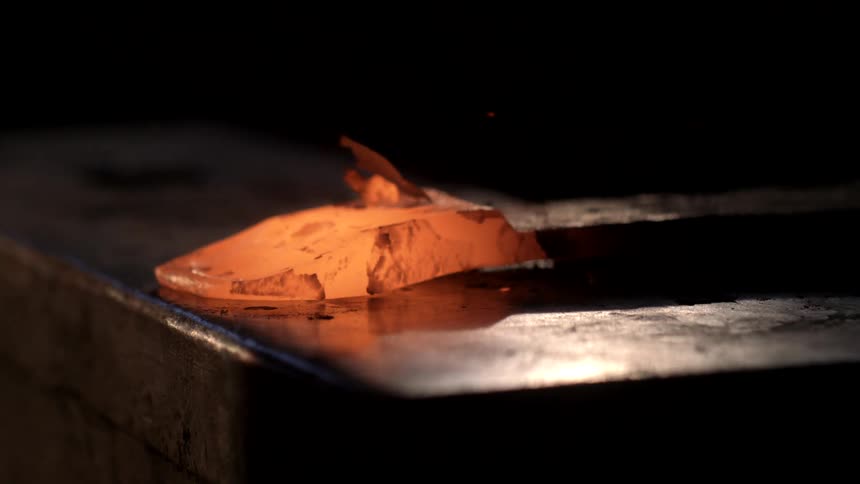 A glowing hot piece of metal is struck with a hammer on an anvil sending sparks and fragments flying in a blacksmithing process