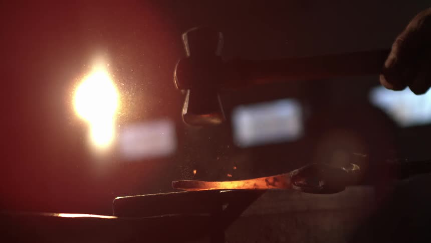 A blacksmiths hand holding a hammer striking a glowing hot piece of metal on an anvil with sparks flying and a bright light source illuminating the scene