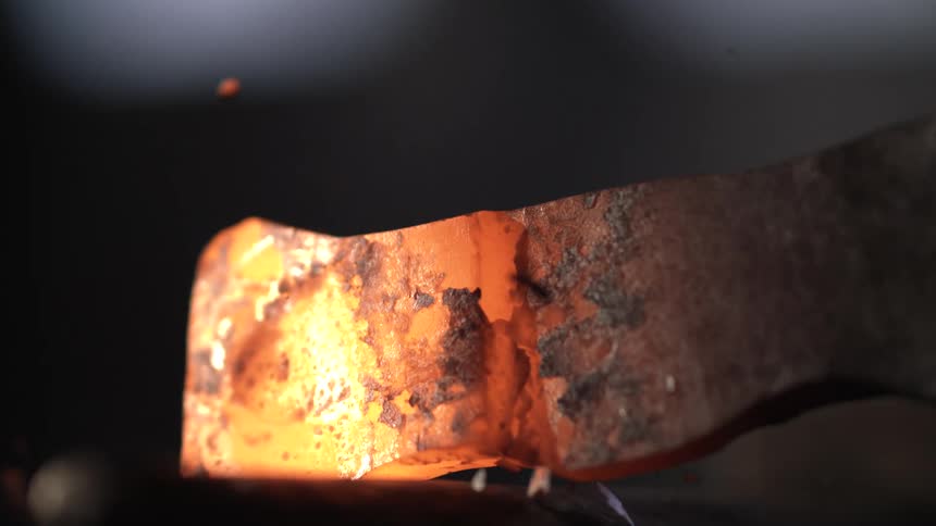 A blacksmiths hand holding a hammer striking a glowing hot piece of metal on an anvil with sparks flying and a bright light source illuminating the scene