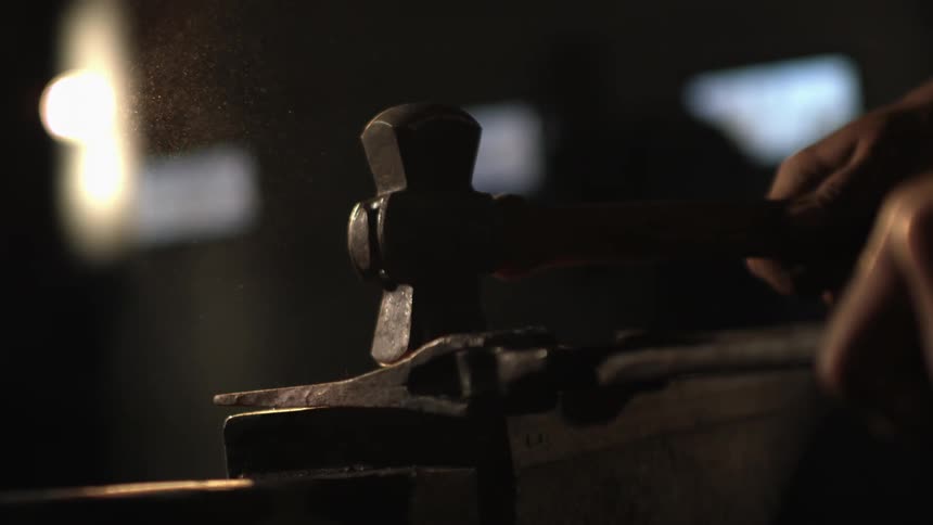 A blacksmith strikes a glowing hot piece of metal on an anvil with a hammer in slow motion causing sparks to fly
