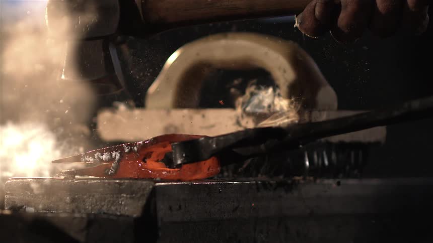 A hammer strikes a glowing redhot piece of metal held by tongs with sparks flying
