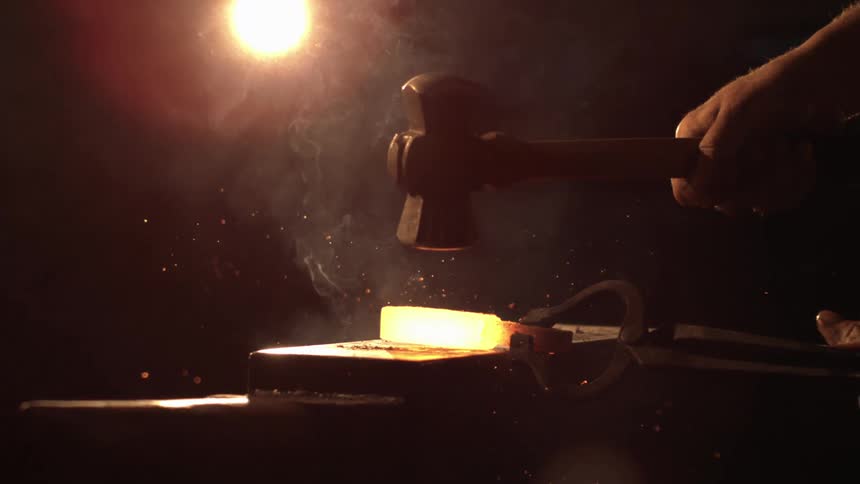 A blacksmiths hands work a glowing hot piece of metal with a hammer on an anvil sending sparks and smoke into the air under a bright light