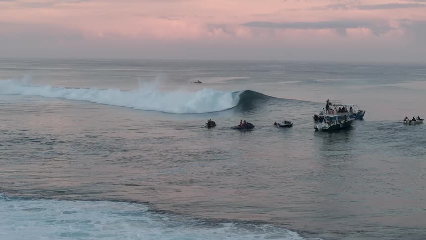 Large waves crash in the ocean under a cloudy sky with a hint of pink with several boats and people visible in the distance suggesting surfers or observers
