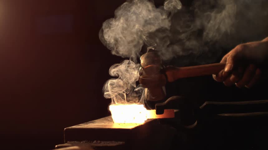 A blacksmith strikes a glowing hot piece of metal with a hammer on an anvil sending smoke rising into the air
