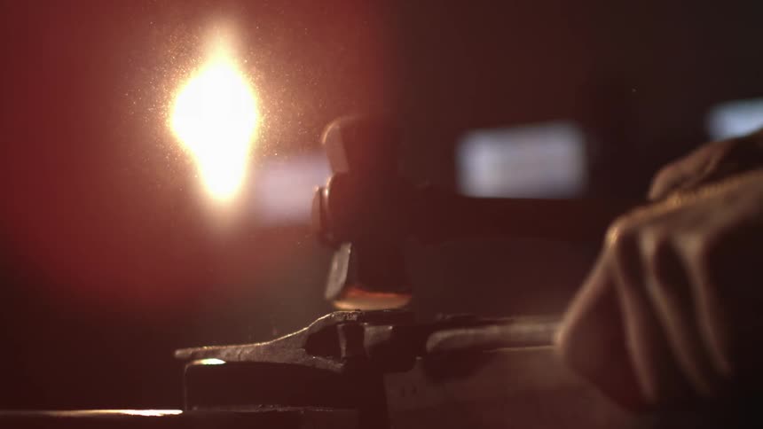 A blacksmiths hand holding a hammer striking a glowing hot piece of metal on an anvil with sparks flying and a bright light source illuminating the scene
